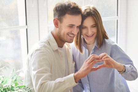 Portrait of cheerful smiling young happy couple making heart gesture from hands by the window at home. Man and woman in love standing indoors. Relationships and Valentines day concept.の写真素材