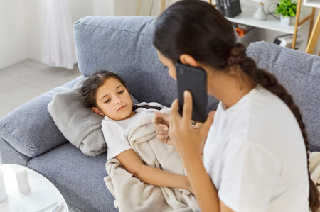 Sick child with mother on sofa. A parent calls the doctor, describing fever and virus illness while offering care, medicine, and rest under a blanket at home. Parental care for sick child.の写真素材