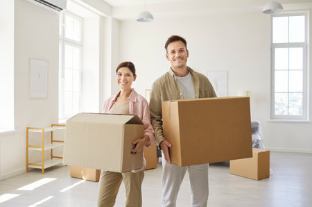 Happy couple moving cardboard boxes into new apartment. Sunlit living room with big windows, minimal...
