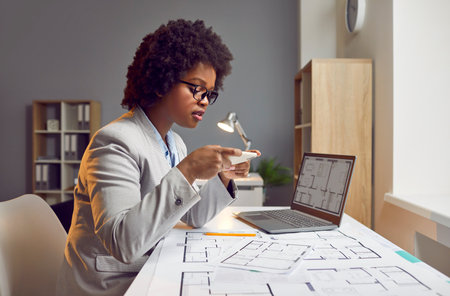 Beautiful young African American woman architect working in her office, sitting at her desk with a notebook PC, holding a modern cellphone and taking photos of house structure layout blueprint plansの写真素材