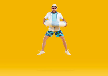 Happy funny man in summer clothes and sunglasses holding inflatable beach ring and jumping on trampoline high in air isolated on orange yellow color studio background. Summer holiday, vacation conceptの写真素材