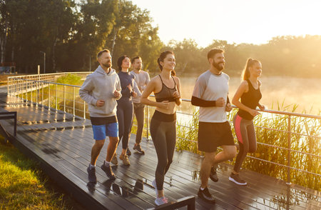 Running friends jogging as group in summer park by lake. Active runners in sportswear training on bridge track, team enjoying morning exercise, teamwork and fresh air with high motivation and energyの写真素材