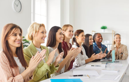 Group of professional women sitting at long table, clapping hands with bright smiles. Happy multiracial female team participating in conference, seminar or corporate training session in modern office.の写真素材
