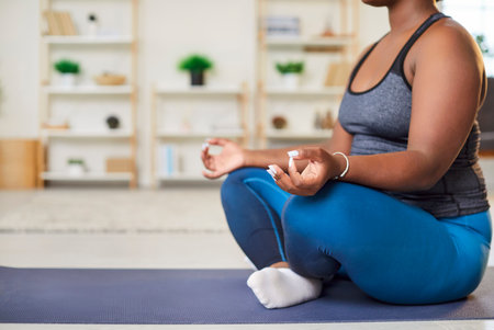 Cropped shot of overweight young African American woman relaxing in Lotus pose, meditating at home. Side view of plus size girl in sportswear doing yoga exercise sitting on floor. Meditation concept.の写真素材