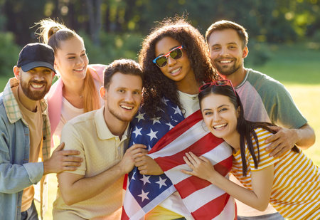 Patriotic cheerful friends posing together, wrapped in USA flag, celebrate America Independence Day, standing in park, enjoying sunny day. Group of mixed race people enjoying celebrationの写真素材
