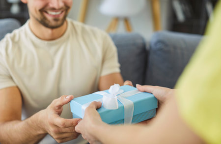 Close-up cropped photo of female surprises her partner with small gift box in cozy, modern living space, hands him present, congratulate with life event, birthday, special occasions or anniversaryの写真素材