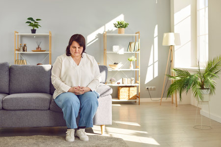 Stressed overweight woman sitting on sofa in living room at home. Troubled plus size woman looking lonely while sitting on couch. Sad fat woman feeling tired and depressedの写真素材