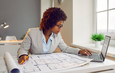 Busy woman architect working with blueprints in her office. Young African American lady sitting at her desk, looking at house plans and layouts and using her laptop computer. Architecture conceptの写真素材