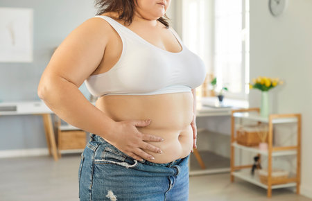 Close up cropped view of plus size woman standing at home, holding her waist with her hands. Overweight female touching stomach, showing body insecurity, health concerns and focus on weight loss.の写真素材