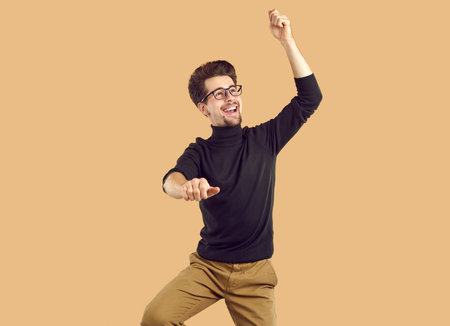 Happy attractive young man is dancing, having fun. Bearded funny guy with glasses wearing black turtleneck and brown trousers on isolated beige background in the studio.の写真素材