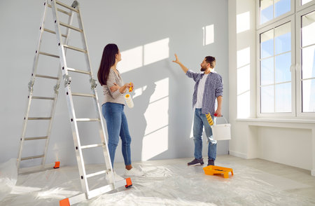 Couple decorating room and painting walls together in their new home. Happy husband and wife with paint rollers renovating their house. Couple standing and discussing color of wallsの写真素材