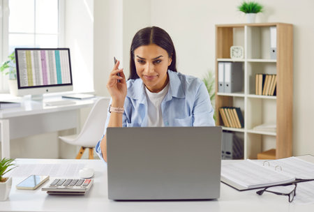 Portrait of a concentrated young business woman accountant sitting at the desk on her workplace and working on a laptop computer with tables and charts at modern office or at home.の写真素材
