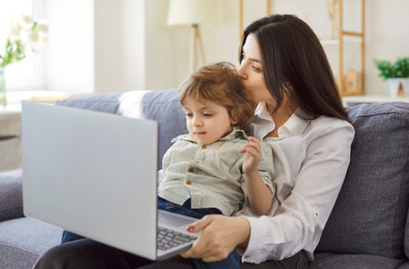 Mother and child with laptop on sofa. A caring woman parent works remotely at home, comforting her toddler kid while multitasking emails and family needs. Balanced remote work, bonding, and family.の写真素材