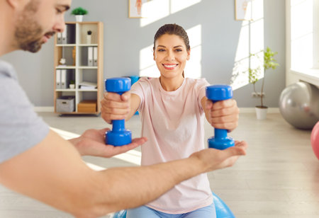 Young happy smiling woman patient doing sport exercise using dumbbells with support from male physiotherapist doctor in rehab clinic. Physiotherapy and physical rehabilitation concept.の写真素材
