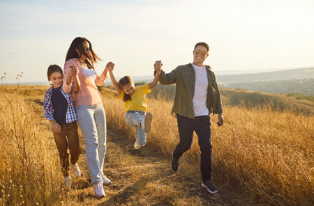 Happy family walking along path through golden meadow, enjoying nature at sunset and fun time together. Parents and children laugh, father and mother holding daughters hands while girl jumpingの写真素材