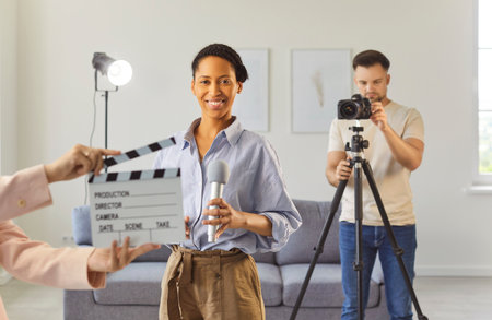 Filming interview or media content in studio, smiling woman holding microphone looking at camera, clapperboard, man operating video camera on tripod, creating video collaborative productionの写真素材