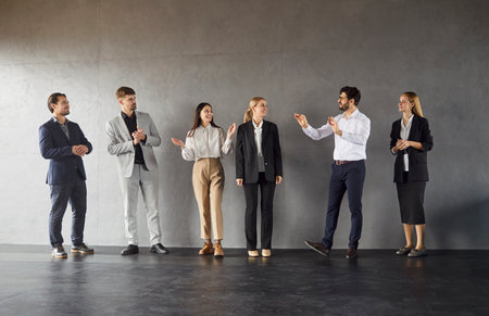 Group of happy confident company employees and colleagues standing in a row on dark gray wall background. Full length portrait of business people men and women in formal clothes talking indoors.の写真素材