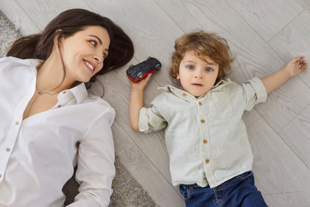 Mother and child play with toy at home. The toddler boy lies on the floor by parent with a toy car,...