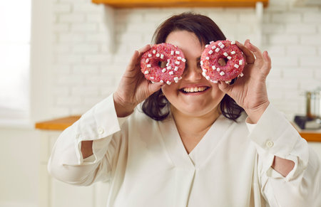Close-up portrait of a happy smiling plump fat plus size brunette woman in white clothes having fun with pink donut cakes on her eyes in the kitchen.の写真素材