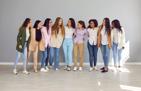 Full size group of a cheerfull happy young diverse women friends or coworkers and company employees looking at each other hugging and smiling together standing on a grey wall background.の写真素材