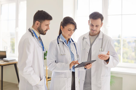 Portrait of a team of young cheerful doctors in white medical uniform looking at tablet screen...