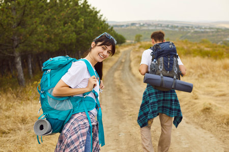 Couple of happy tourists walking on trail by pine forest with backpacks. Young woman turned around...