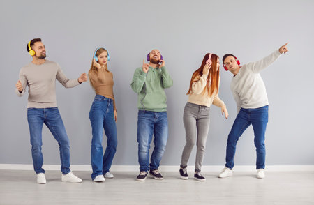 Full length portrait of a young joyful happy people friends or students men and women wearing casual clothes having fun listening to music in headphones standing in a row on a gray wall background.の写真素材