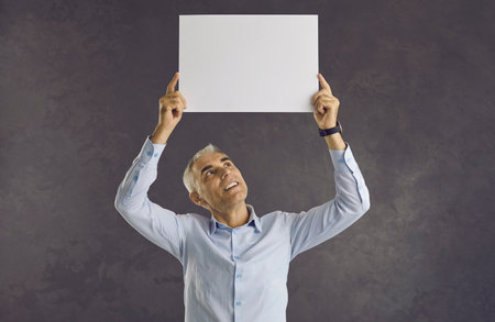 Caucasian senior man looks at white blank sheet of paper he holds over his head. Mature businessman standing on gray background makes announcement, votes or expresses his position.の写真素材