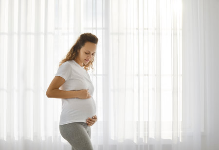 Happy young pregnant woman stand near window at home touch big belly. Smiling healthy Caucasian...
