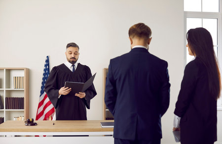 Serious fair male American judge in gown uniform enforces punishment on young man. Lawyer and defendant standing and listening to judge giving judgment, back, rear view. Court trial, law in US conceptの写真素材