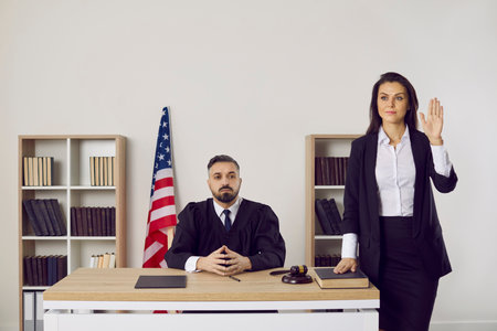 Young male judge sit at desk in court, female witness oath on constitution before trial. Man juror or prosecutor against American flag in law. Justice and jurisdiction concept.の写真素材