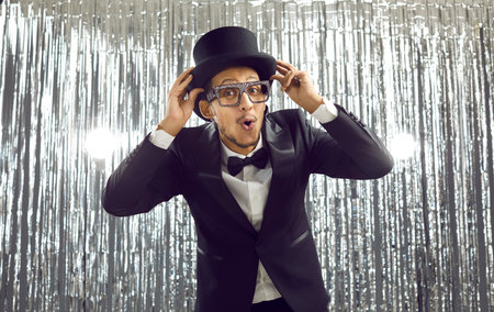 Cheerful stylish dark-skinned young man in elegant black suit having fun at party. Guy in cool glasses is holding his hat and with funny expression looks at camera standing on shiny silver background.の写真素材