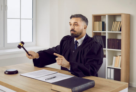 Portrait of young judge in robe sitting at his table in courtroom, hitting sound block with gavel, asking court for silence or giving judgment at the end of trial session. Law and justice conceptの写真素材