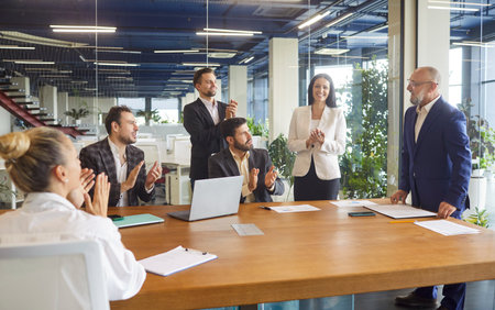 Business people applaud happy speaker after meeting presentation in office modern boardroom. Excited corporate staff sitting and standing at desk to clap and celebrate success with ovationの写真素材