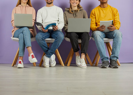 Young people use laptops and notebooks to prepare for lectures, exams and interviews. Legs of multiracial young students sitting in row on chairs on purple wall background. Low section.の写真素材