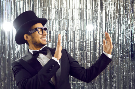 Smiling young latin american in black tuxedo holding in the hands of empty space on shiny silver background. Positive elegant guy in glasses, classic suit, bow tie and top hat looks at the copy space.の写真素材