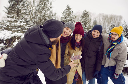 Group of cheerful young friends talking while walking outdoors in snow in winter forest. Young men...