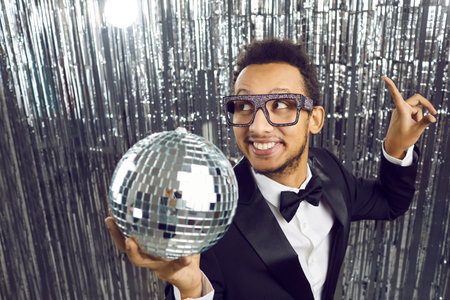 Lets get party. Cheerful young dark-skinned disco man in suit with shiny silver mirror disco ball in his hand. Close up of happy businessman on silver party background. Concept of corporate partyの写真素材