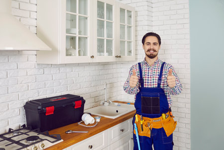 Happy handsome mustached young man in uniform overalls standing near the sink in a modern kitchen, showing thumbs up and smilingの写真素材