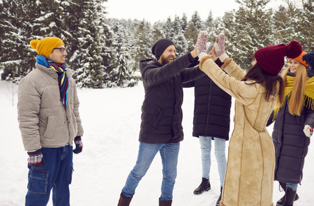 Playful men and women having fun in winter woods. Best friends surrounded by other friends give each other high five while walking on winter walk. Group of friends have fun together on winter weekend.の写真素材