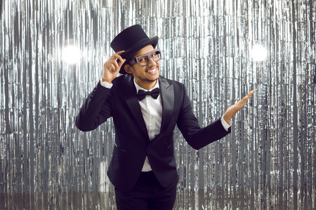 Portrait of positive young dark-skinned man who politely greets you holding on to his top hat. Funny cheerful man in elegant tuxedo stands on background of shiny foil party curtains. Banner.の写真素材