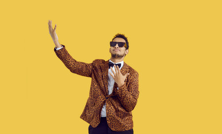 Happy man wearing a leopard party suit, bowtie and cool sunglasses dancing in the studio. Funny young guy listening to music and dancing isolated on a yellow colour backgroundの写真素材