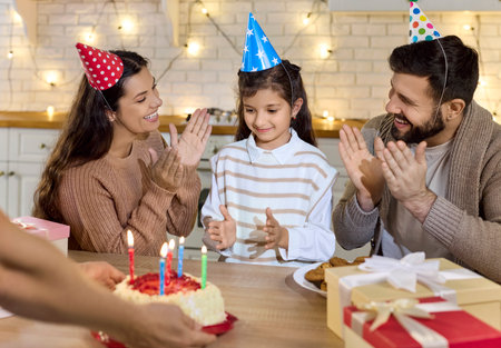Family birthday party with daughter, cake, candles. At home, smiling parents watch as the child greets a lit cake and wrapped gifts on the table. Warm family celebration of childhood at home.の写真素材