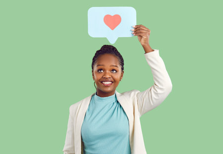 Smiling young black woman isolated on green studio background hold speech bubble with painted heart. Happy African American girl with talk balloon show love and care. Charity and volunteer.の写真素材
