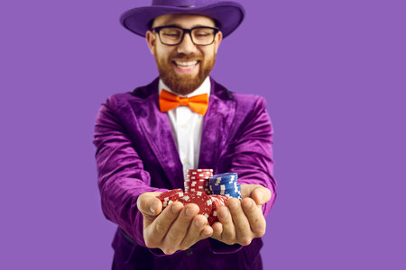 Close up of poker chips in hands of joyful and satisfied man isolated on purple background. Man in velvet purple jacket, bow tie and hat is smiling and looking at poker or casino chips in his hands.の写真素材
