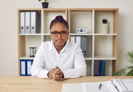 Young African American business woman in white shirt and eyeglasses sitting at office table and looking at camera with serious face expression. Portrait of businesswoman at online web conferenceの写真素材