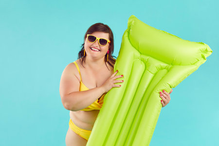 Portrait of happy, cheerful, smiling woman in yellow bikini and sunglasses standing isolated on blue background and holding green inflatable mattress. Summer holiday at beach conceptの写真素材