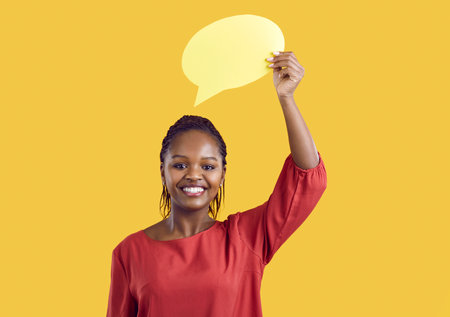 Portrait of smiling African American woman isolated on yellow studio background show mockup speech bubble. Happy African girl demonstrate empty talk balloon with copy space for ad.の写真素材