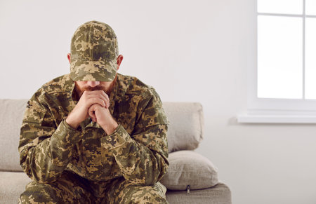 Serious anxious and nervous military man suffers from depression while sitting alone at home. Pensive man in military uniform sitting on sofa propping his head with his hands. PTSD concept.の写真素材