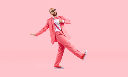 Cheerful, funny and humorous young man in pink suit takes step on pastel pink background. Stylish cool guy in suit with suspenders, bow tie and sneakers makes step looking into camera. Full length.の写真素材